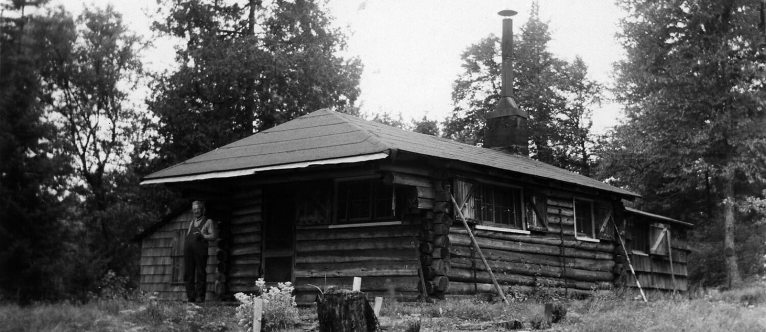 Black and white photo of a man standing outside a log cabin